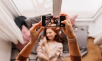 Photo of hands holding phone during photoshoot. Ginger girl posing for her friend at home.