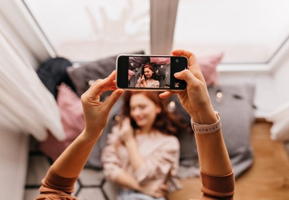 Photo of hands holding phone during photoshoot. Ginger girl posing for her friend at home.